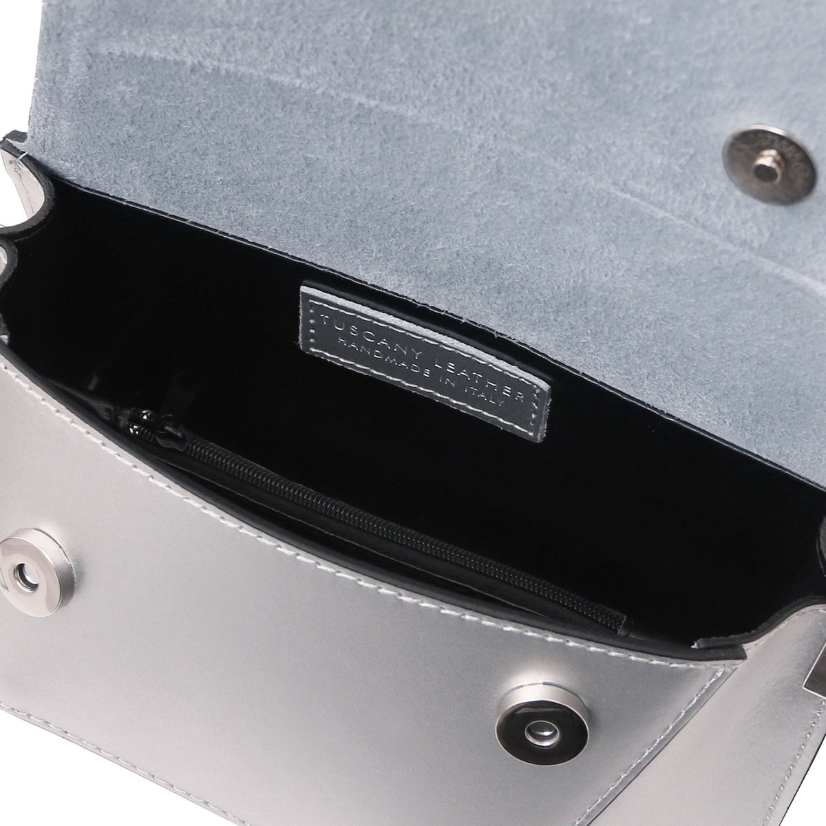 Close-up of an open silver metallic handbag showing a black interior, zippered compartment, and a silver-tone brand label that reads TUSCANY LEATHER HANDMADE IN ITALY.