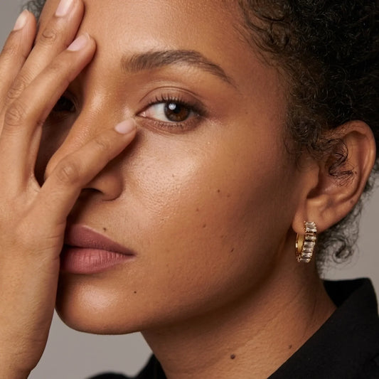 Close-up of a woman wearing a gold huggie hoop earring adorned with small clear stones, with her hand covering part of her face.