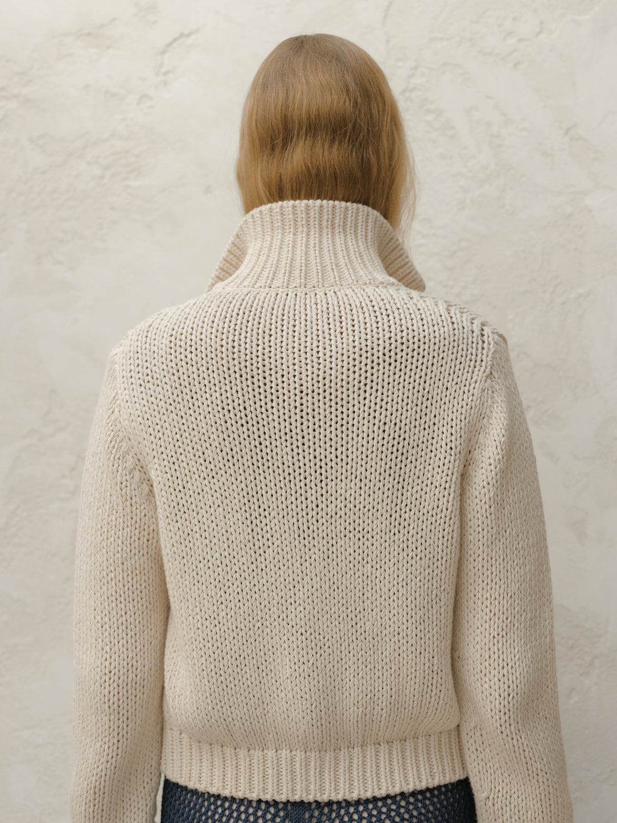 Back view of a model wearing a cream chunky knit bomber jacket with a ribbed hem against a textured light wall.
