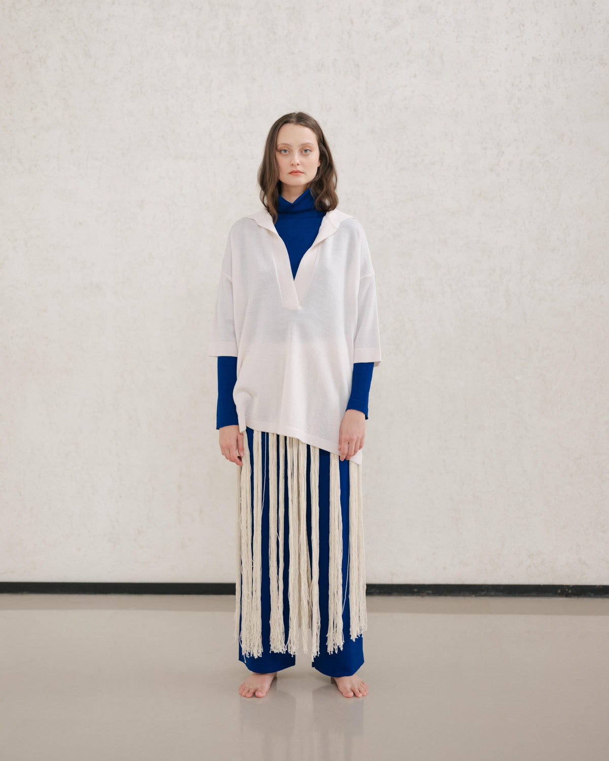 Model wears a pale cream Maxi polo en laine mérinos extrafine with a royal blue underlayer and a long fringe skirt.