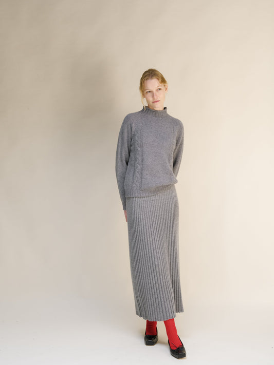 Full-length shot of a woman in a gray cable knit sweater and matching ribbed maxi skirt against a beige backdrop.