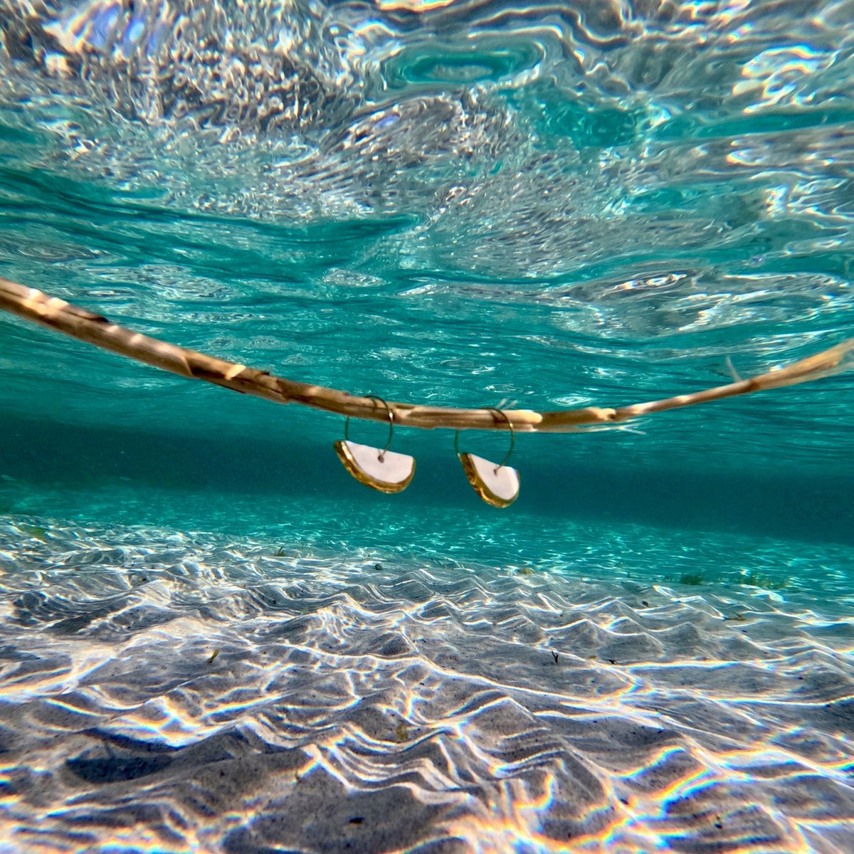 Ceramic earrings with gilded edges floating in clear blue water with a sandy ocean floor.