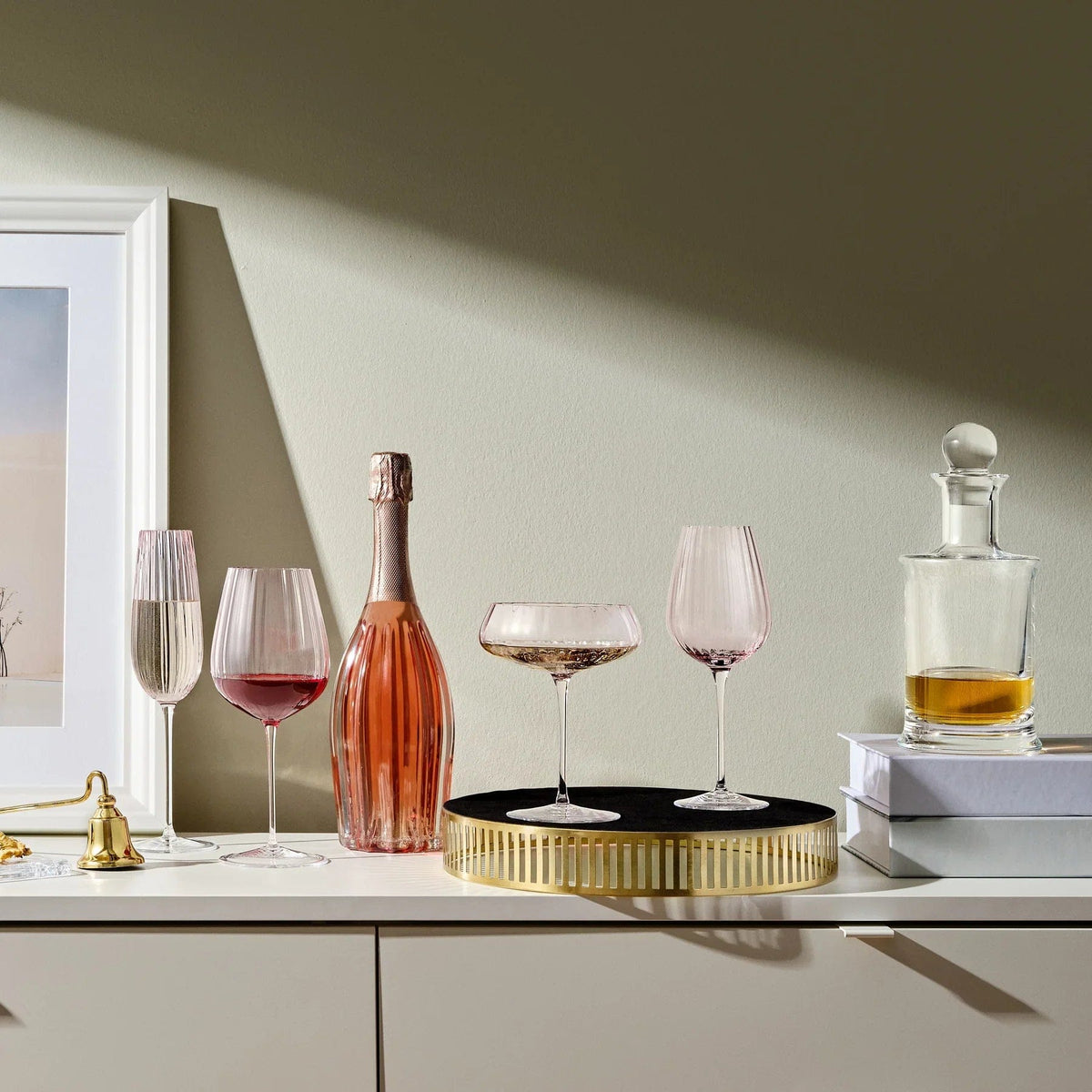 Dusty rose glassware arrangement with a pink bottle on a white surface