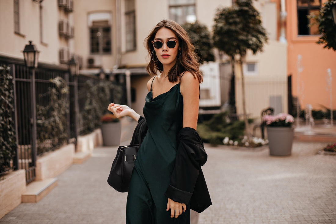 Stilvolle Frau in einem dunkelgrünen Trägerkleid und Sonnenbrille geht mit einer schwarzen Lederhandtasche durch eine Stadtstrasse.