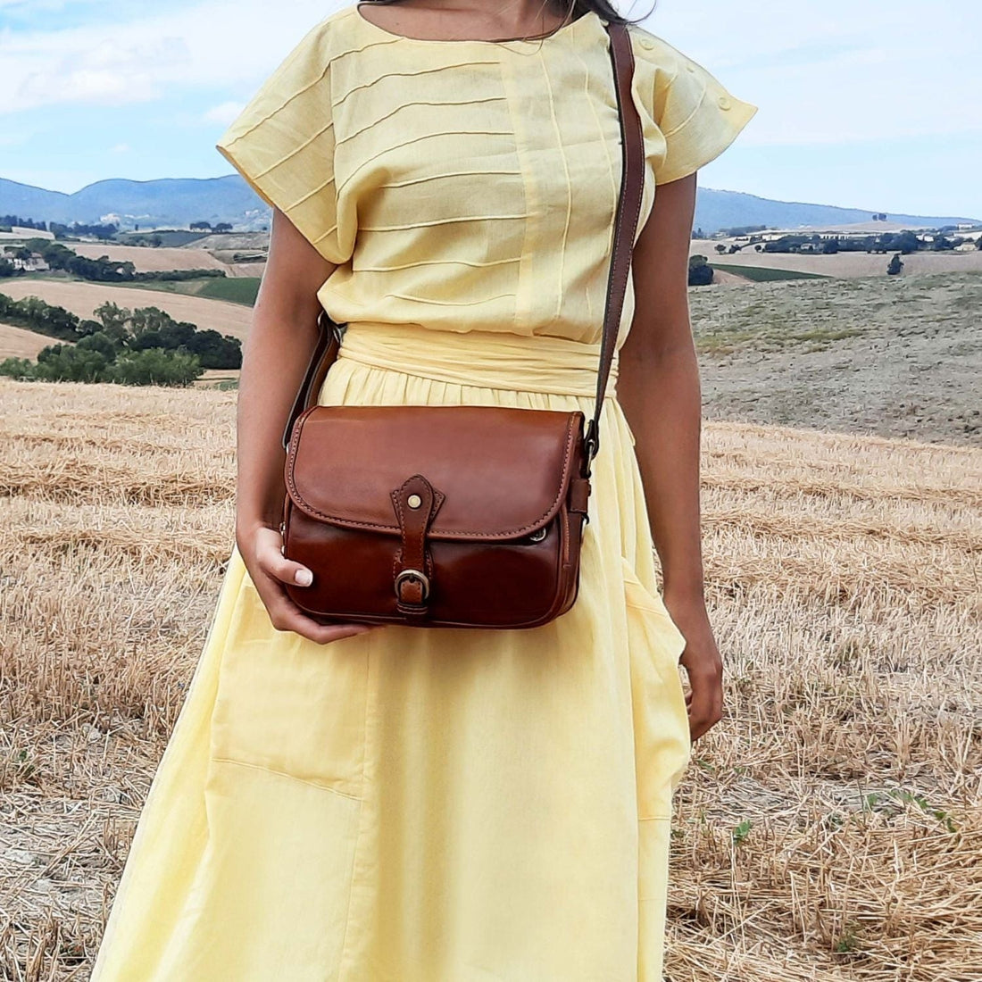 Mulher com vestido amarelo-claro usa uma mala a tiracolo em pele castanha num campo soalheiro.