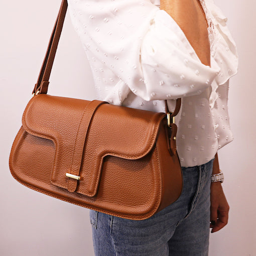 Sac à bandoulière en cuir grainé marron avec rabat avant, porté par une personne vêtue d’un chemisier blanc à pois.