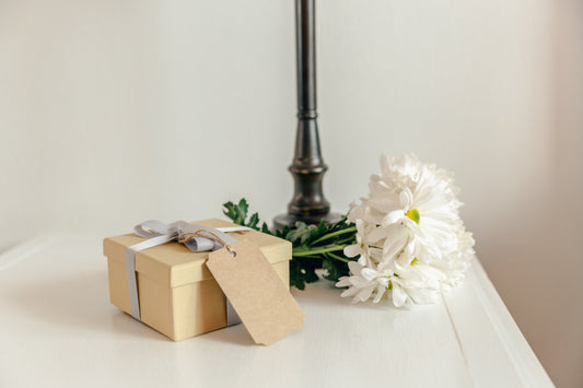 Caja de regalo beige con cinta gris y etiqueta en blanco junto a un ramo blanco sobre una superficie blanca