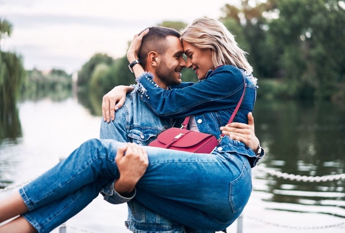 Romantic couple in denim embrace by a lake with a bright pink handbag.