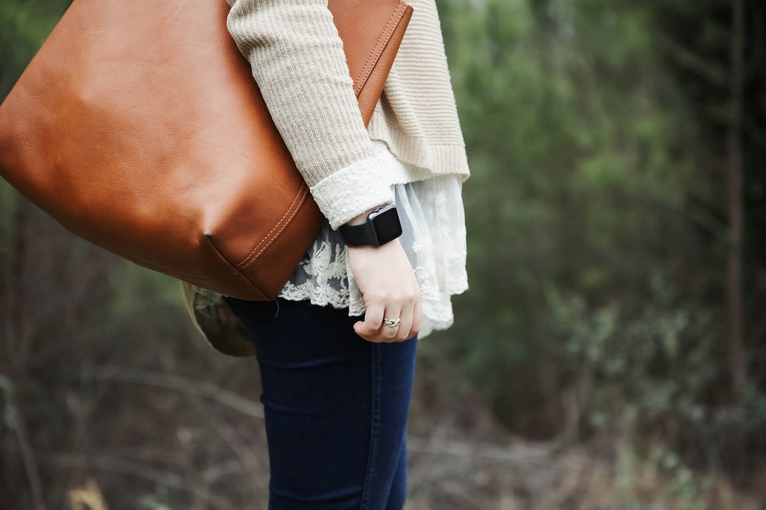Tan leather tote slung over a shoulder of a person in a cream knit top and lace skirt outdoors among trees.