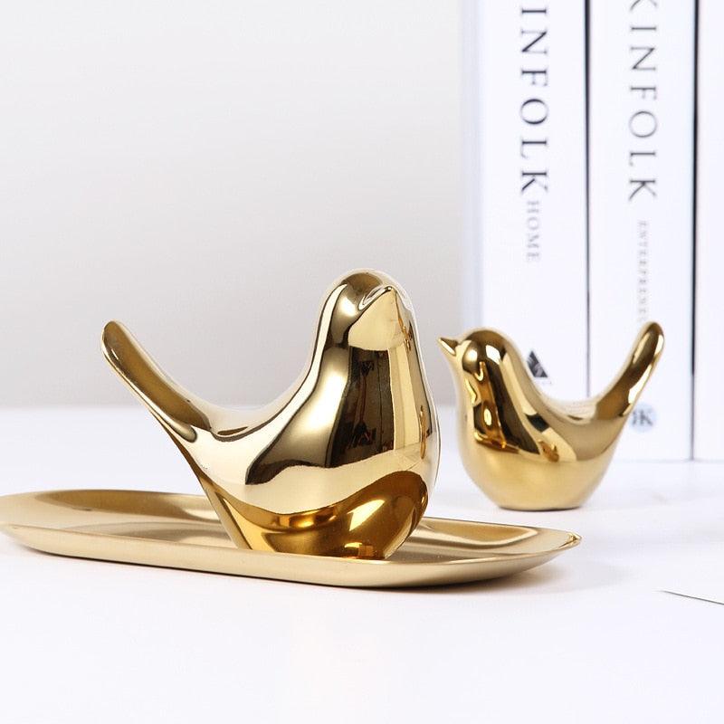 Sculture dorate a forma di uccello su un vassoio riflettente, davanti a uno sfondo bianco con dorsi di libri sullo sfondo