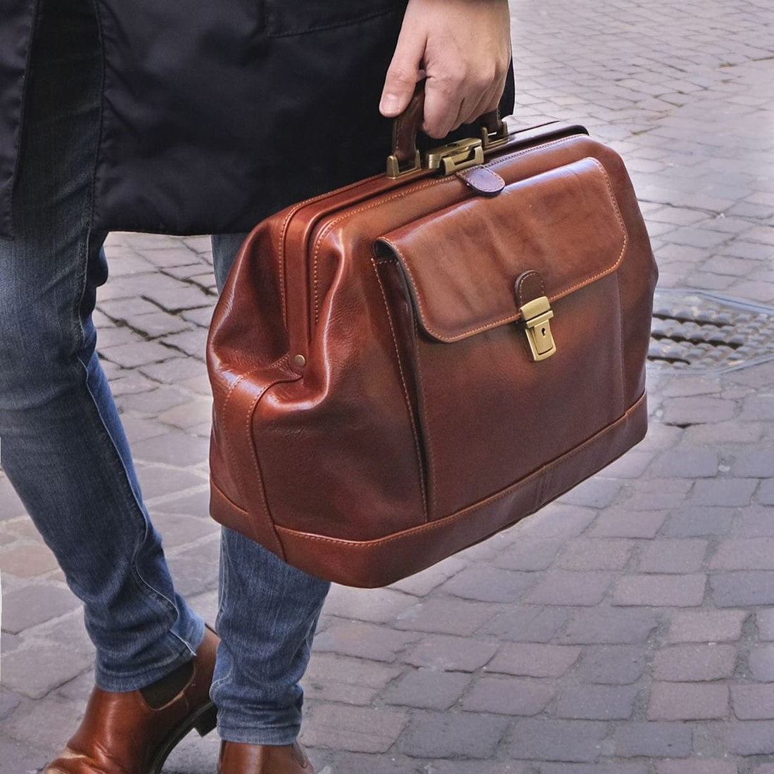 Personne en jean portant une serviette en cuir marron avec finitions en laiton dans une rue pavée.