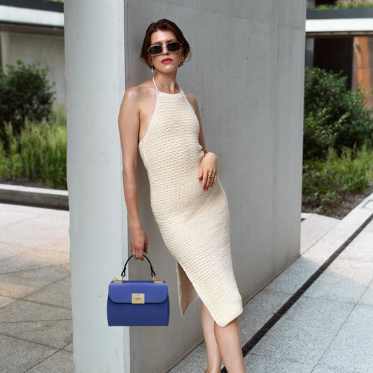 Mulher elegante com vestido halter de malha cor de creme encostada a um pilar de betão, segurando uma mala azul.