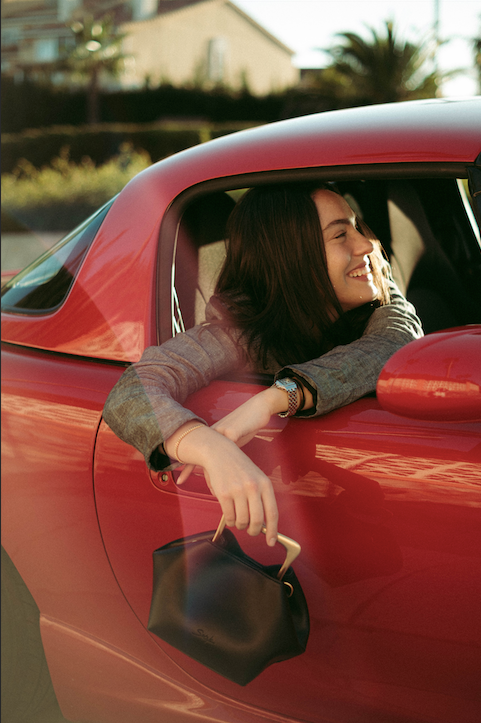 Mujer sonriente asomada a la ventanilla de un coche clásico rojo mientras sostiene un pequeño bolso de hombro de piel marrón oscuro.