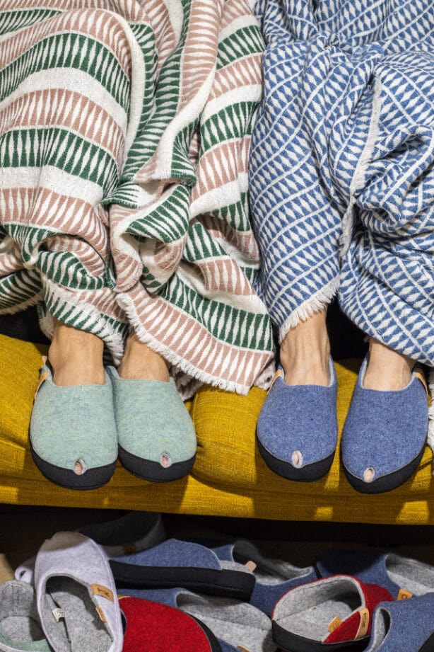 Duas pessoas com chinelos às riscas azuis e verdes estão sentadas com mantas às riscas num sofá amarelo