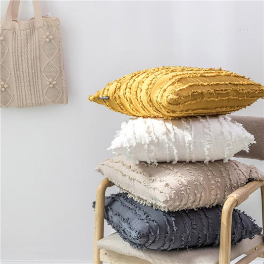 Cojines texturizados apilados en beige, blanco y mostaza sobre una silla de madera, con un bolso tejido colgado de una pared blanca.