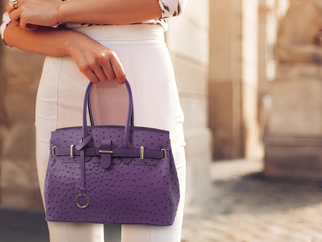 Sac à main violet en cuir embossé façon autruche, porté par une personne en pantalon blanc dans une rue de ville
