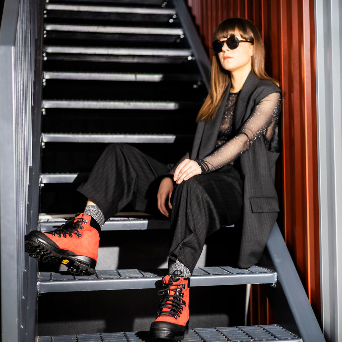 Frau in einem dunklen Anzug mit transparenten Ärmeln sitzt auf einer Metalltreppe und trägt rote Wanderstiefel.