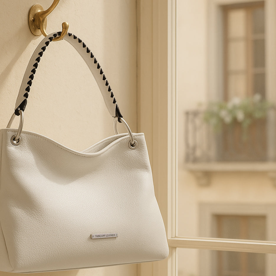 Weisse handgefertigte Lederhandtasche mit geflochtenem Henkel hängt an einem Wandhaken neben einem sonnenbeschienenen Fenster in einer italienischen Wohnung.