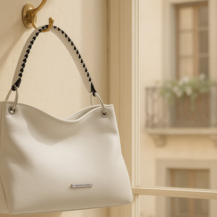 White handcrafted leather handbag with braided handle hanging on a wall hook by a sunlit window in an Italian apartment.