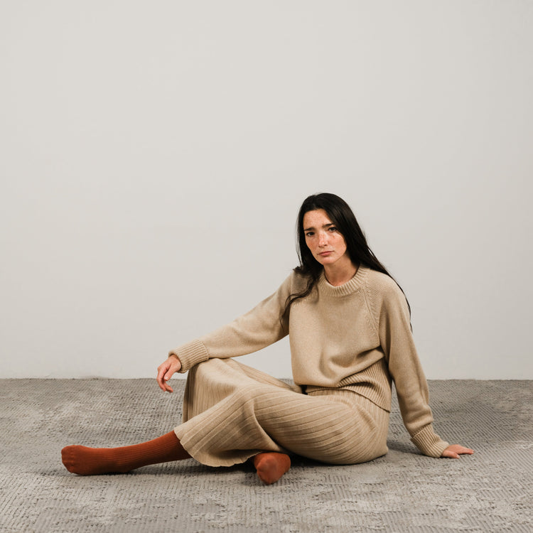 Mulher com conjunto bege em malha de cashmere sentada sobre um tapete texturado diante de uma parede clara e neutra.
