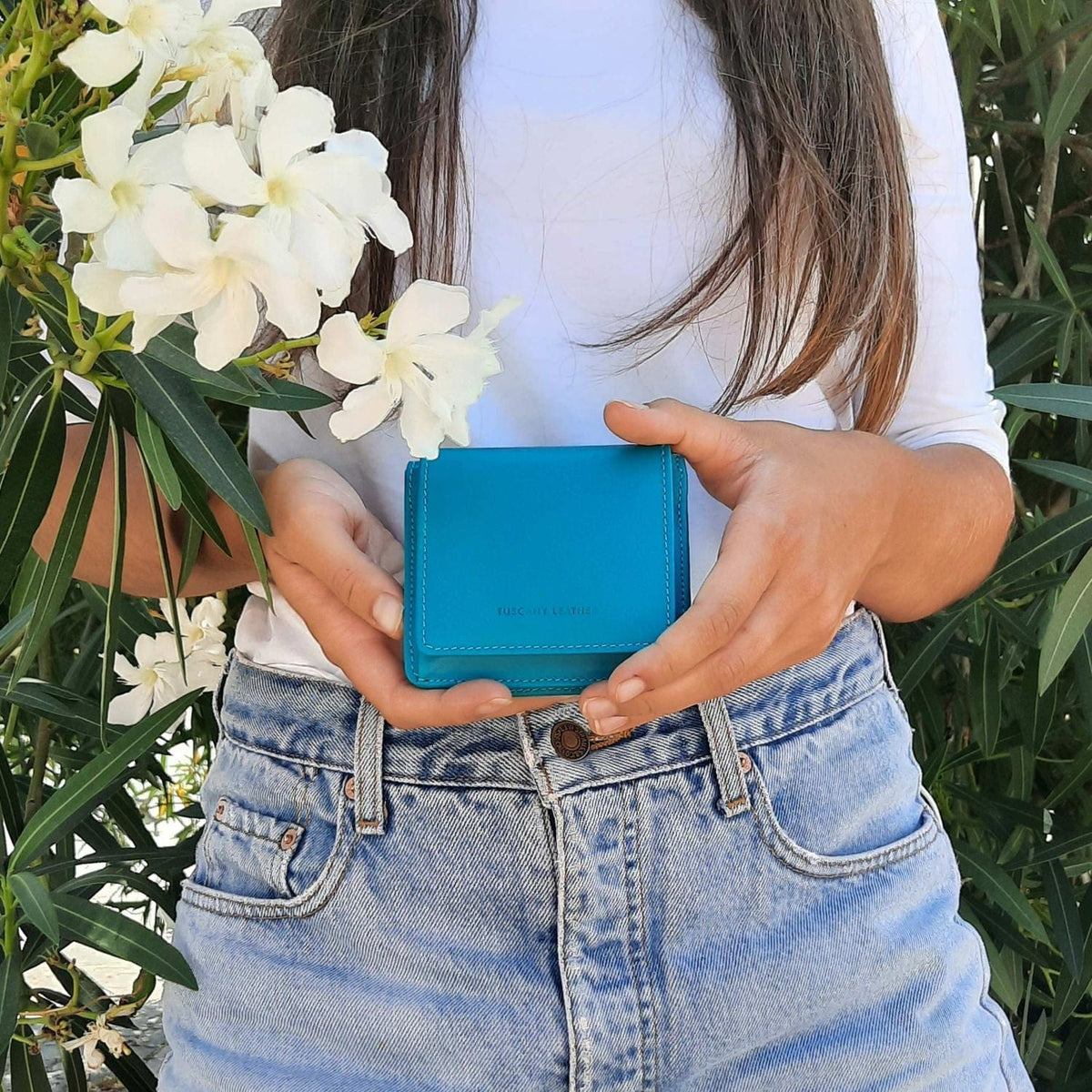 Donna con top bianco e jeans che tiene un portafoglio turchese all’aperto, con fiori bianchi nella scena.