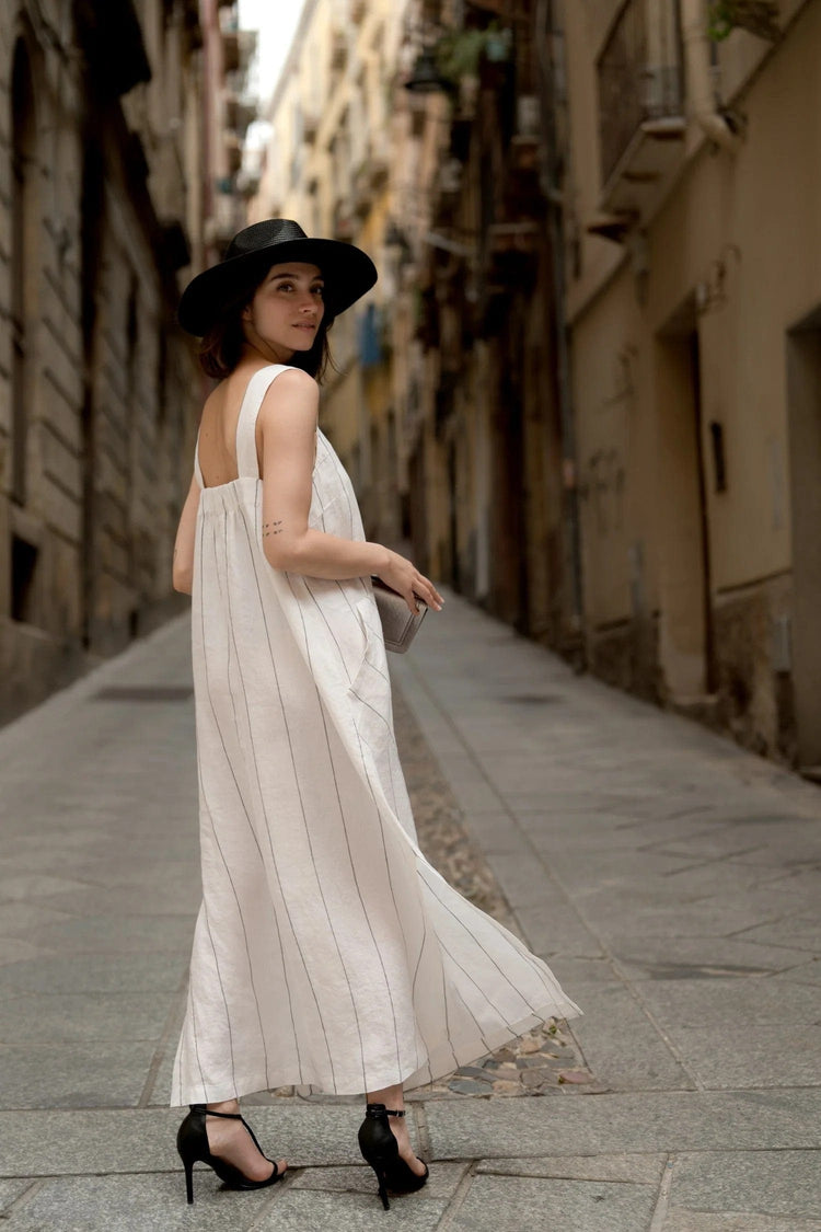 Woman wearing a flowing striped linen summer dress and black hat walking down a narrow cobblestone street in Europe.