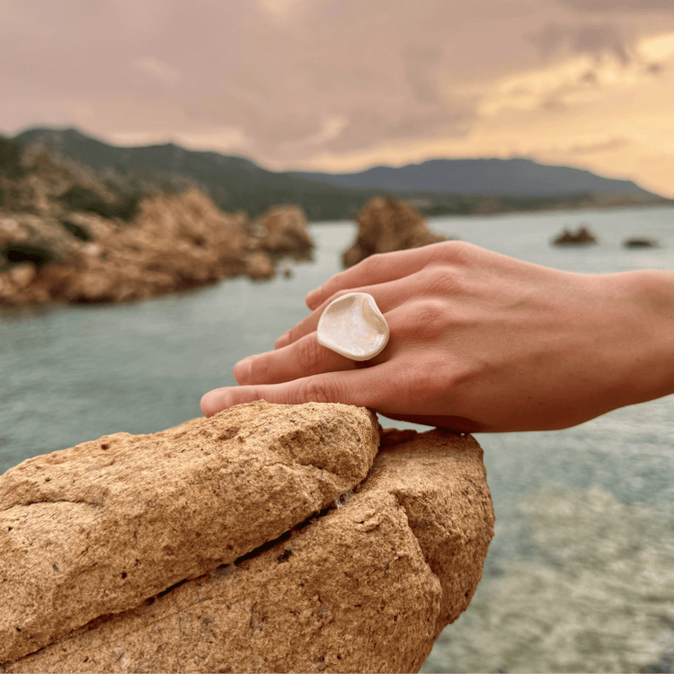 Hand resting on a rough rock by the sea wearing a large iridescent shell ring.