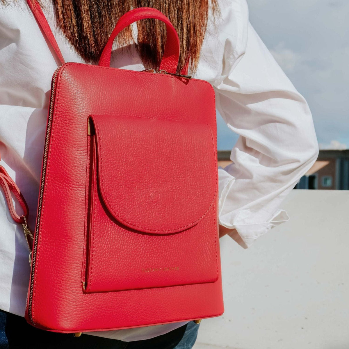 Zaino rosso brillante in pelle con tasca frontale curva e manico superiore, portato da una persona con camicia bianca.