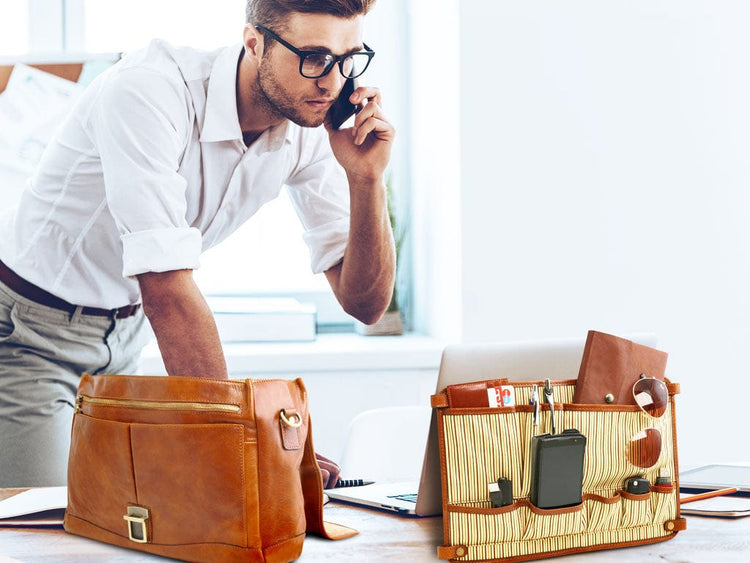 Man in a white shirt and glasses on a phone beside tan leather business bags and a laptop on a desk