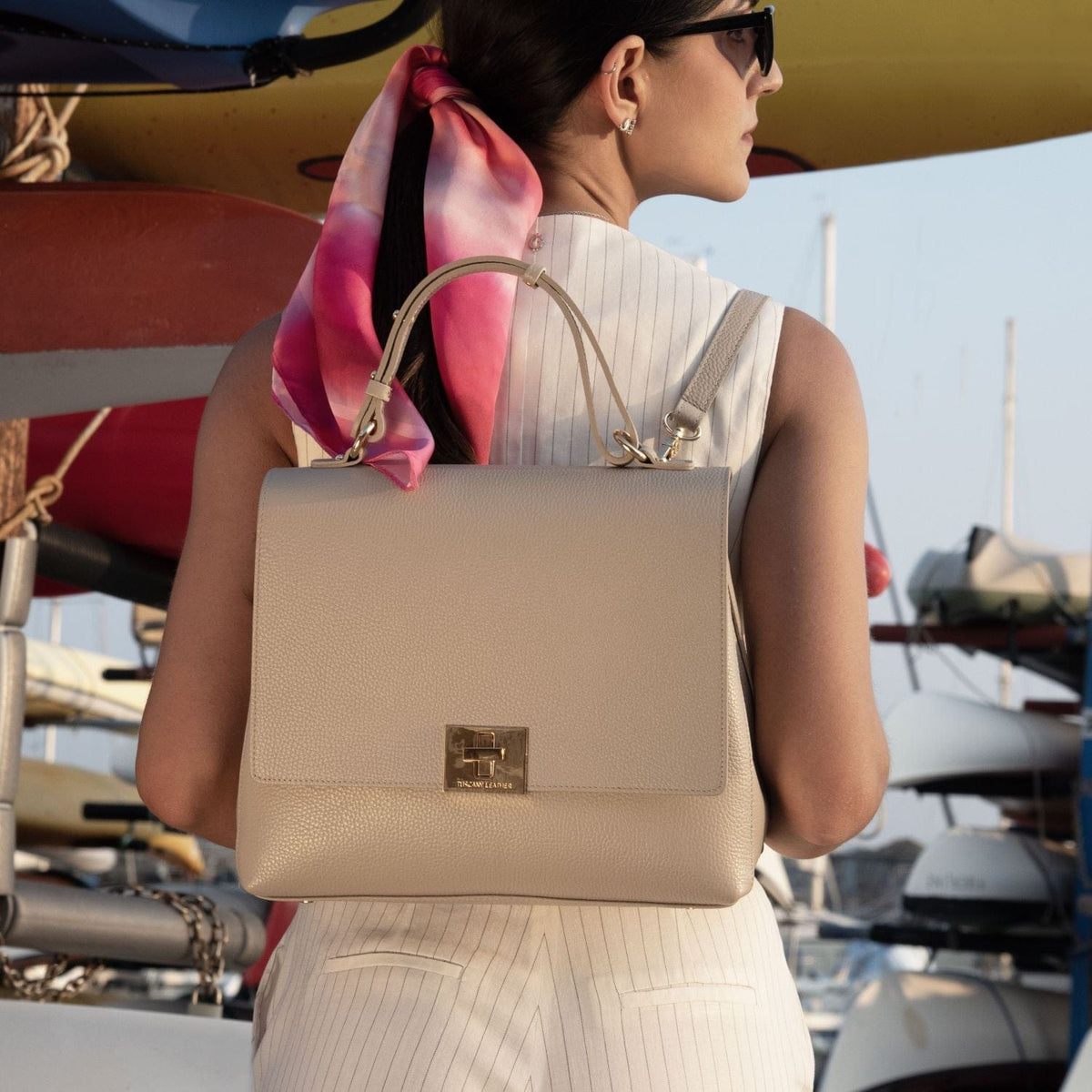 Beige Handtasche aus genarbtem Leder mit goldfarbenen Metallteilen, getragen von einer Frau in der Nähe eines Yachthafens.