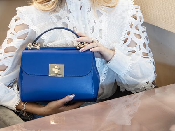 Woman in a white lace blouse holding the Armonia royal blue Italian leather handbag with a gold clasp, handcrafted entirely in Italy, seated at a glossy pink marble table.