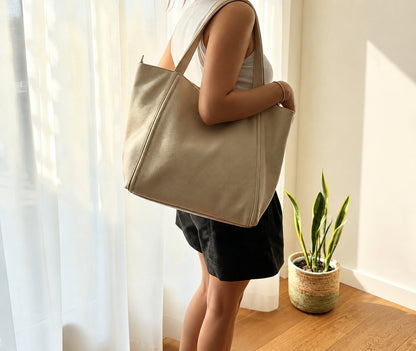 A woman wearing a white tank top and black shorts carries a large beige canvas tote bag over her shoulder in a brightly lit room with sheer curtains, a wooden floor, and a potted snake plant.
