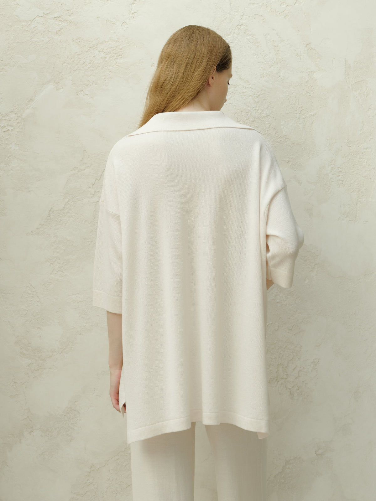 Vue de dos d’un mannequin en Merino Wool Maxi Polo crème pâle au tombé ample devant un mur texturé
