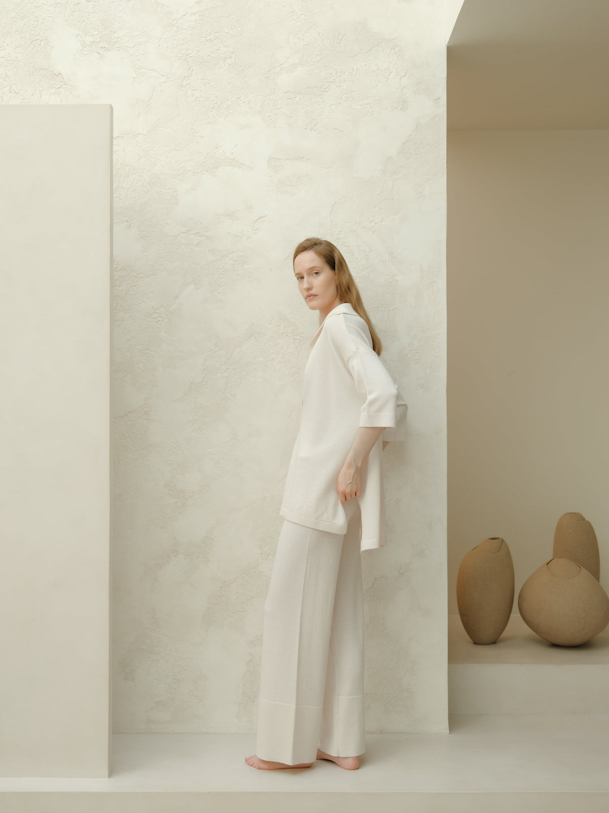 Mannequin portant a white Maxi polo en laine mérinos extrafine and pantalon large assorti stands barefoot against a pale textured wall.