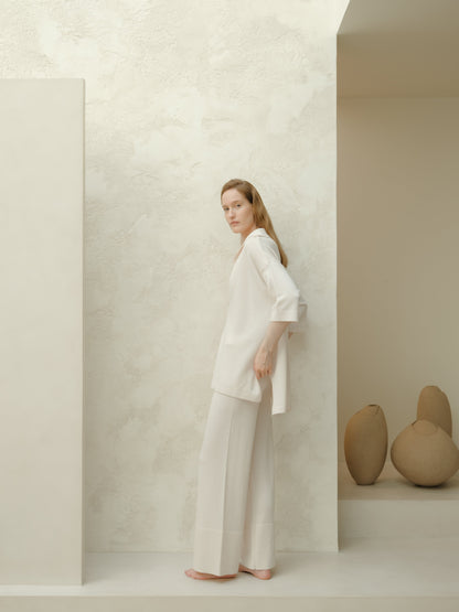 Mannequin portant a white Maxi polo en laine mérinos extrafine and pantalon large assorti stands barefoot against a pale textured wall.