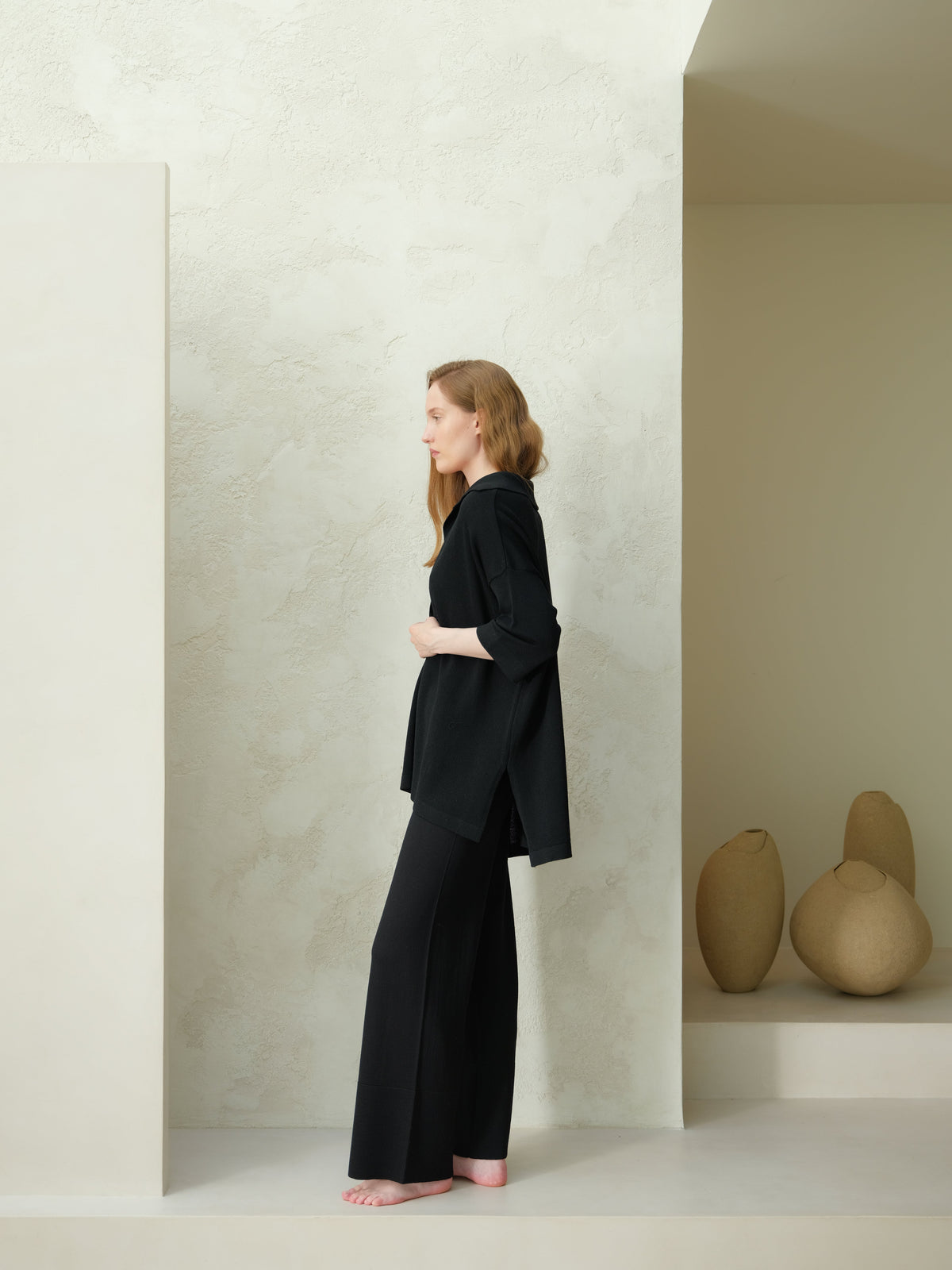 Profile view of a mannequin portant a black Maxi polo en laine mérinos extrafine with wide-leg trousers against a pale textured backdrop.