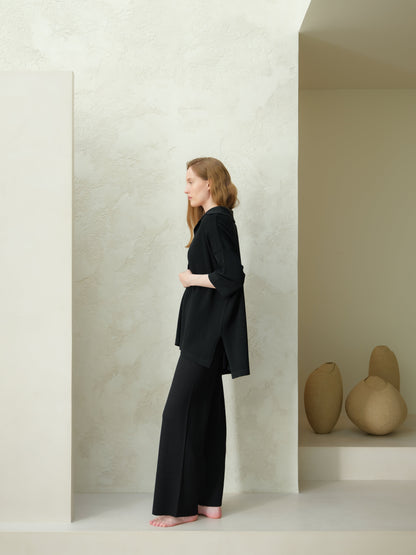 Profile view of a mannequin portant a black Maxi polo en laine mérinos extrafine with wide-leg trousers against a pale textured backdrop.