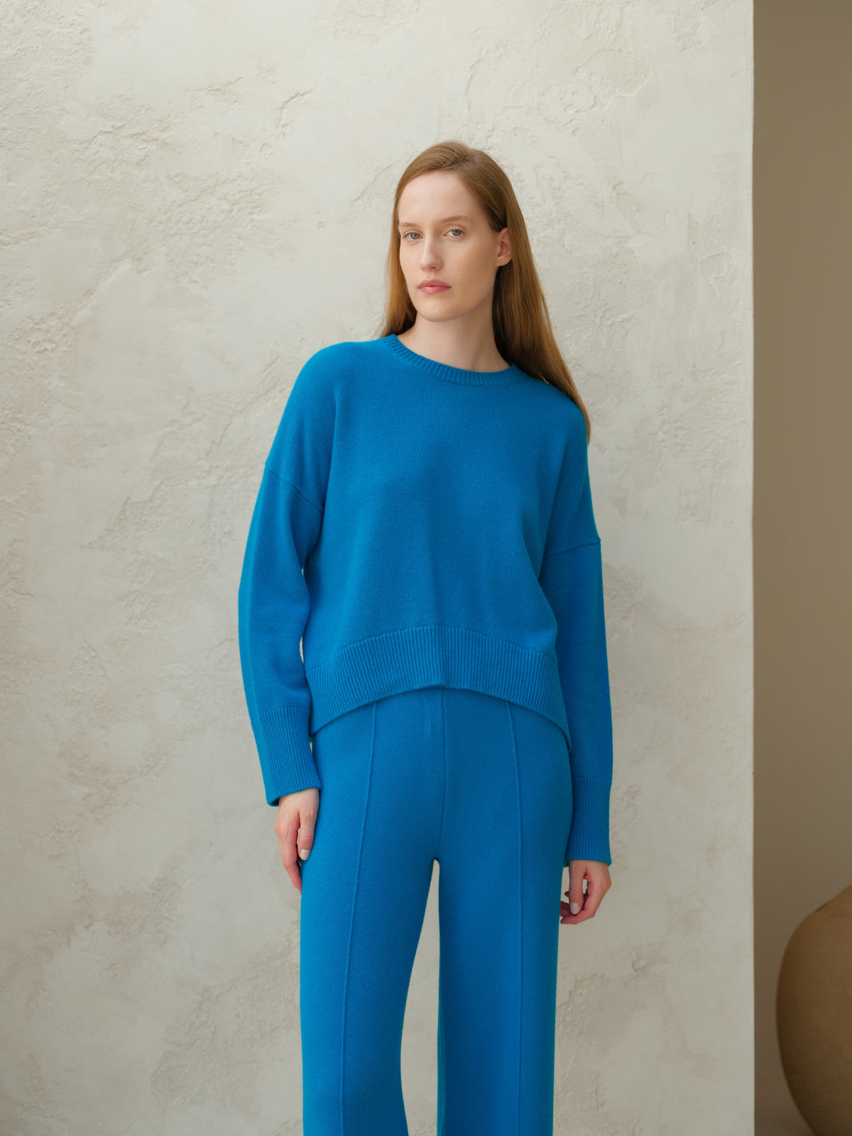 Modella con bright blu cashmere mix maglione girocollo con matching blu knit pants against a light wall.