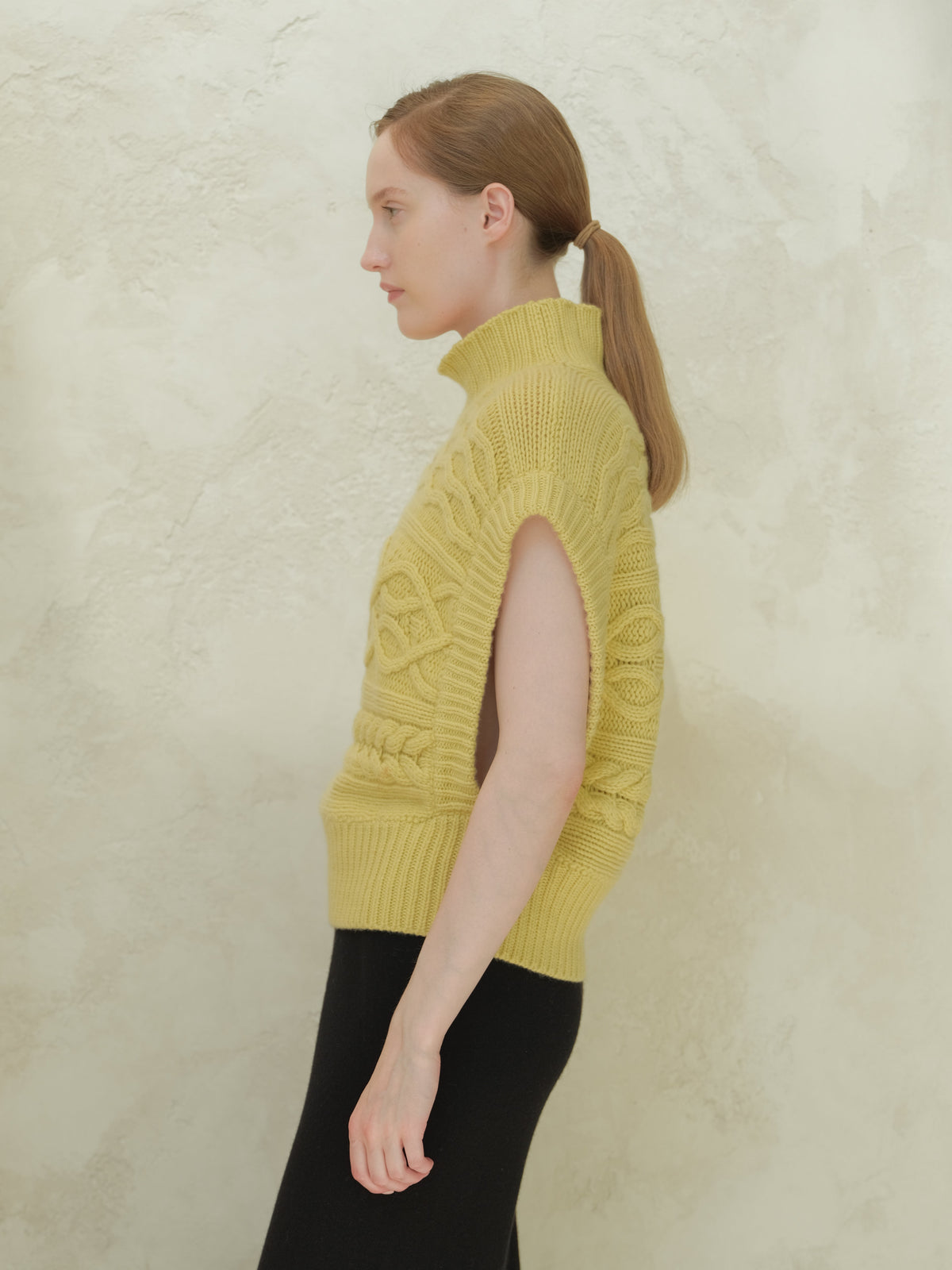 Side view of a modella con a pale yellow collo alto cable knit maxi tunic con sleeveless silhouette.