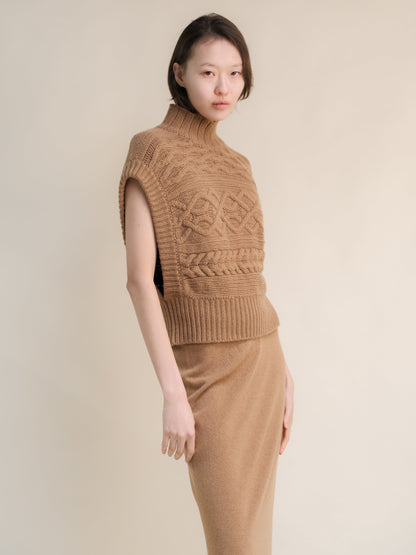 Tunica in maglia beige senza maniche a collo alto con grandi trecce, abbinata a una gonna color cammello