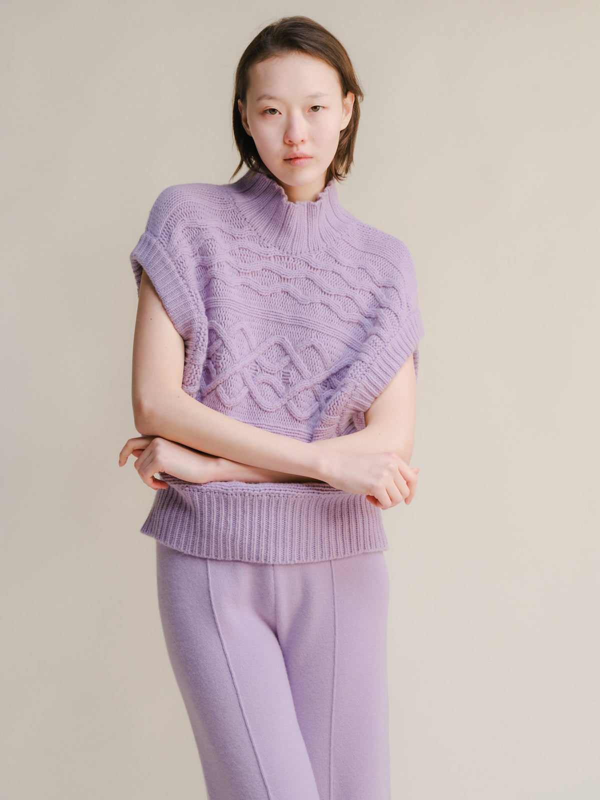 Lilla chunky knit sleeveless tunic con a collo alto e cable pattern worn con matching lilac pants.