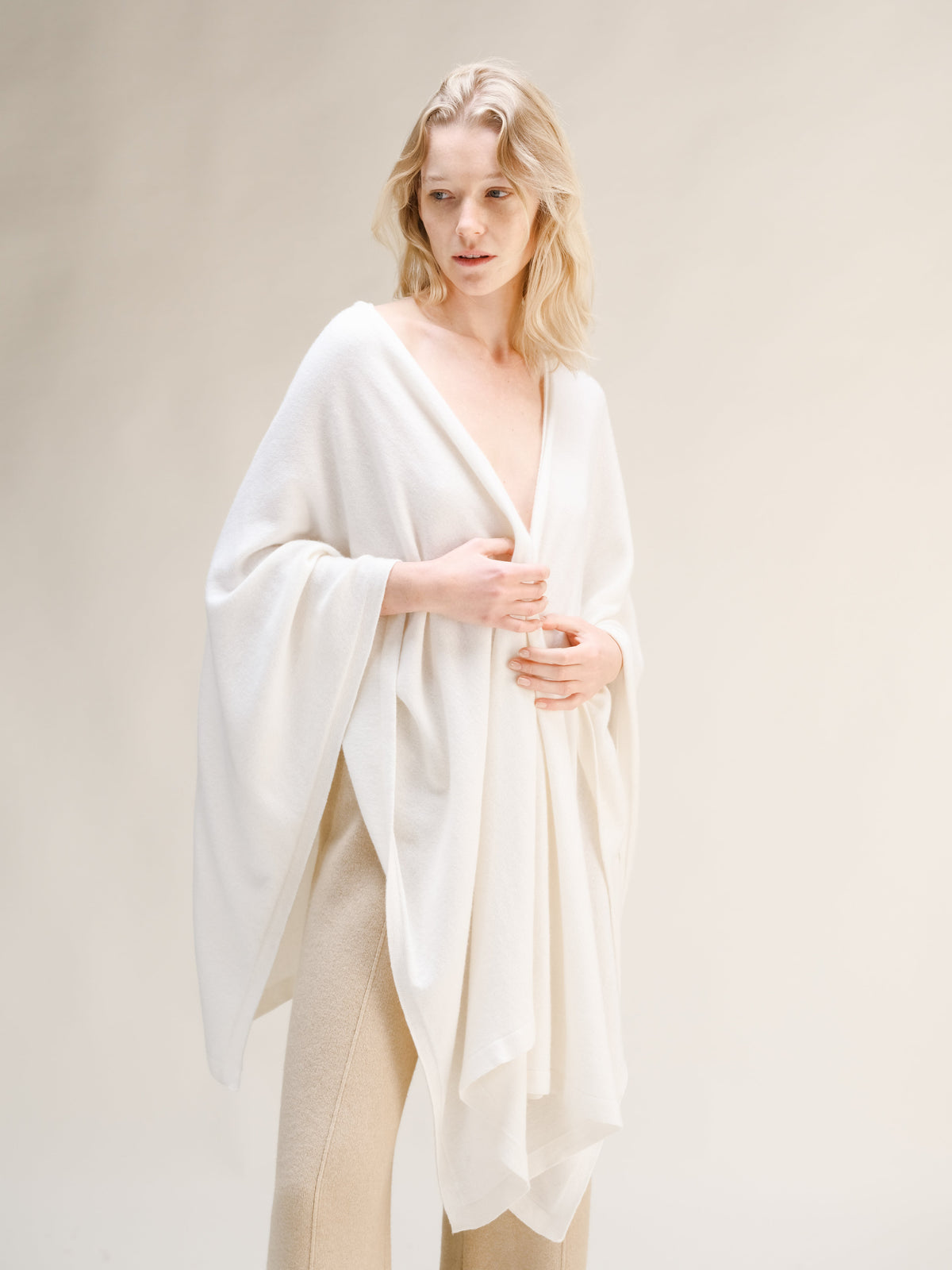 Modella con maxi cappa bianca in misto cashmere drappeggiata sulle spalle e pantaloni in maglia beige