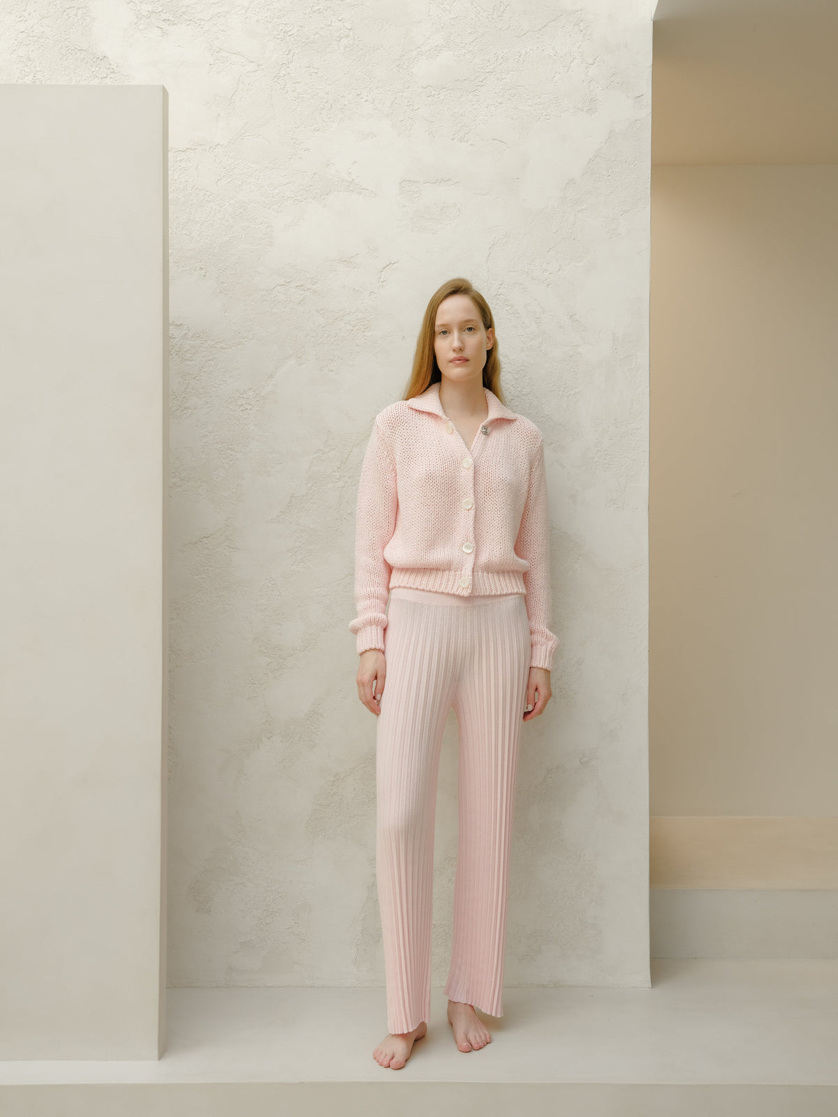 Model wearing pale pink knitted cardigan and matching ribbed pants standing barefoot against a textured pale wall.