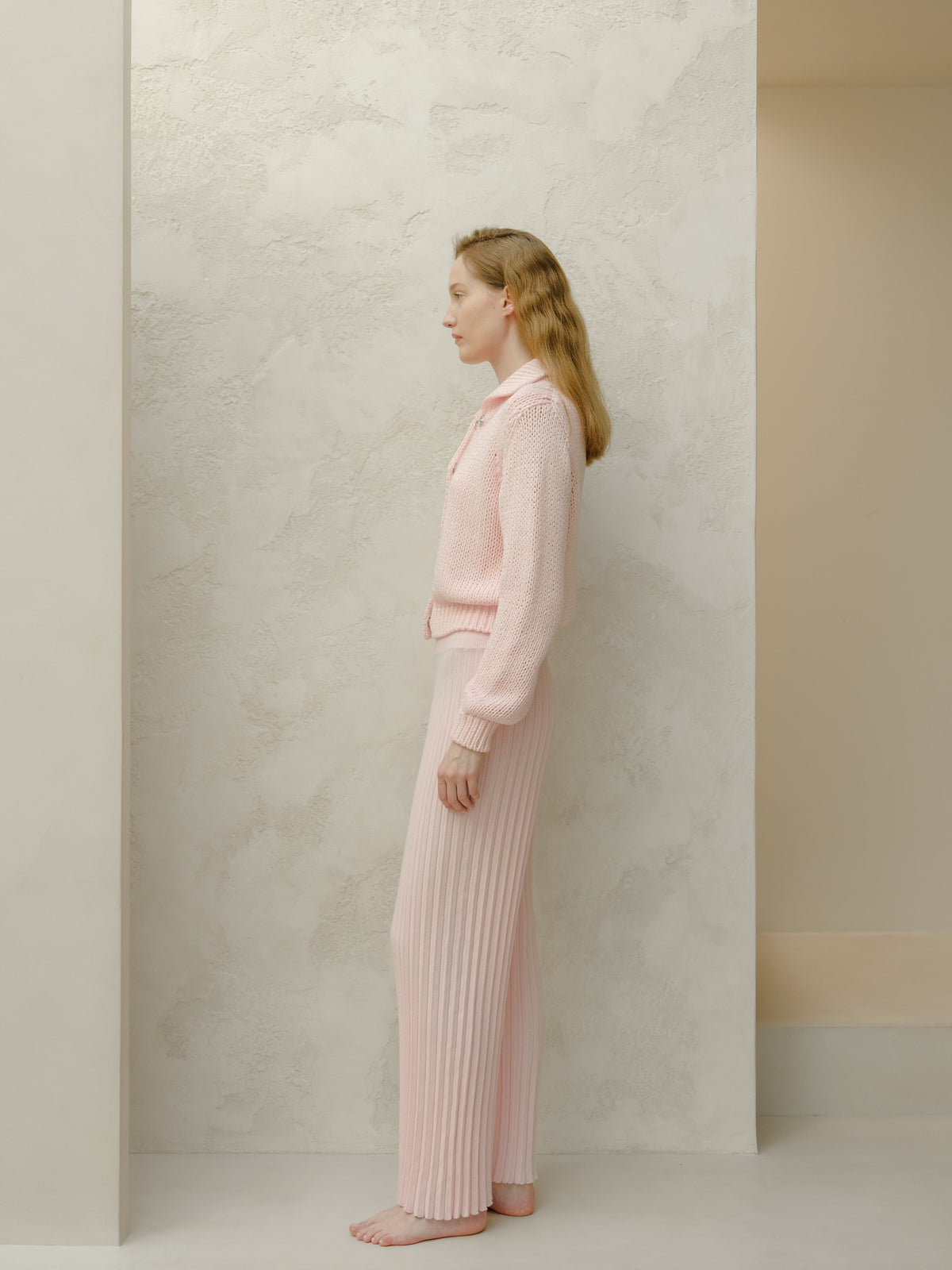 Side view of a model in a pale pink knit set with a sweater and ribbed wide-leg pants against a textured pale wall