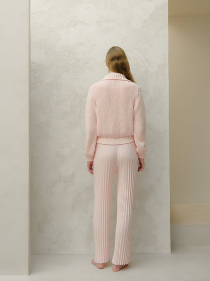 Back view of a model wearing a pale pink knit sweater and matching ribbed pants against a textured wall