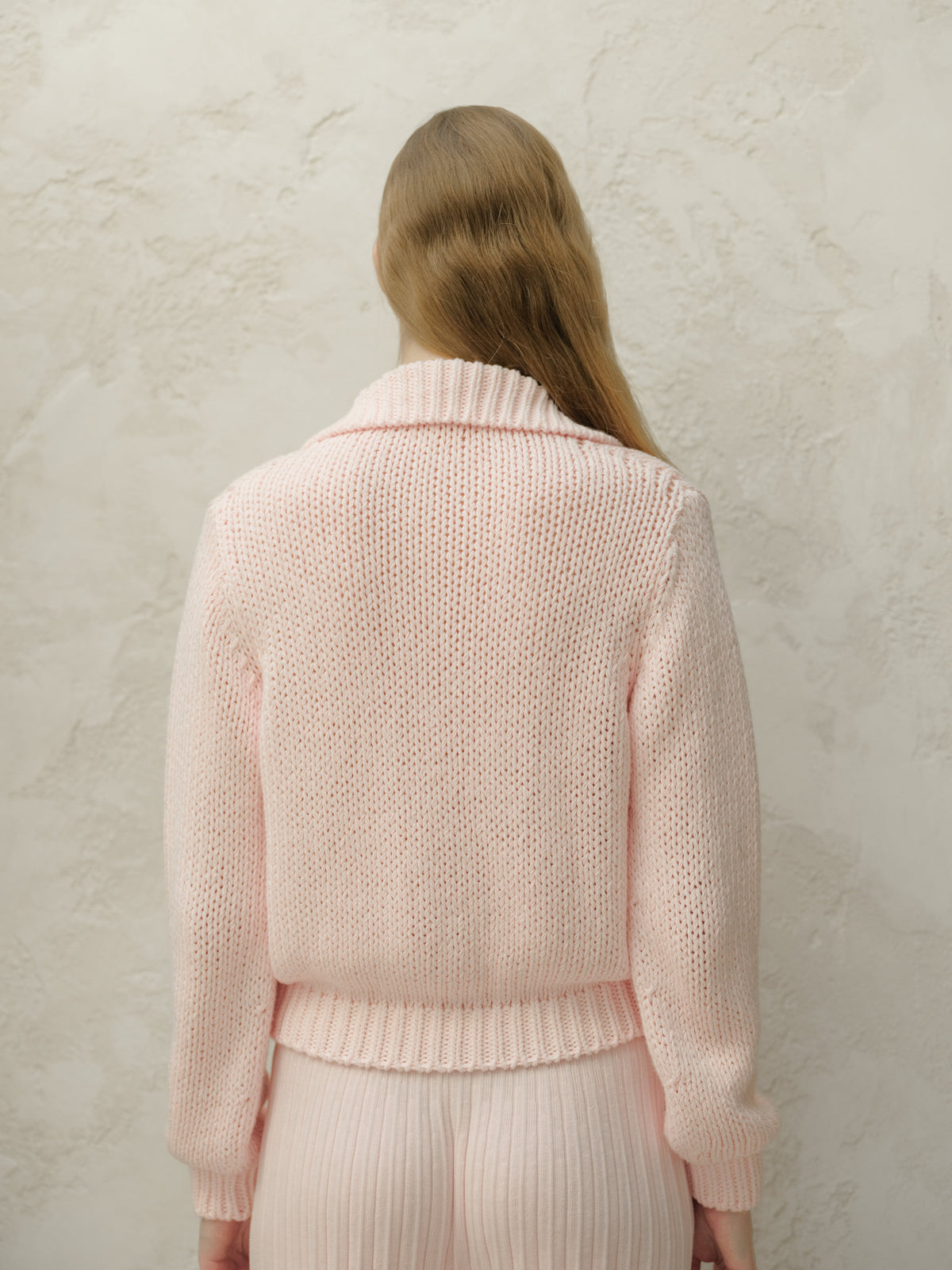 Back view of a model in a soft pink knit cardigan showing relaxed fit and ribbed hem against a textured wall