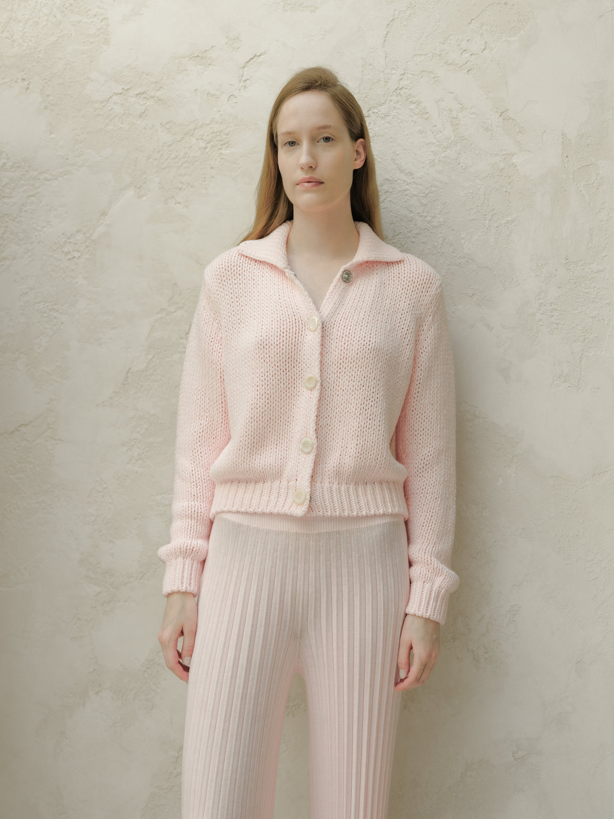 Front view of a model wearing a soft pink knitted cardigan with large white buttons against a textured beige wall