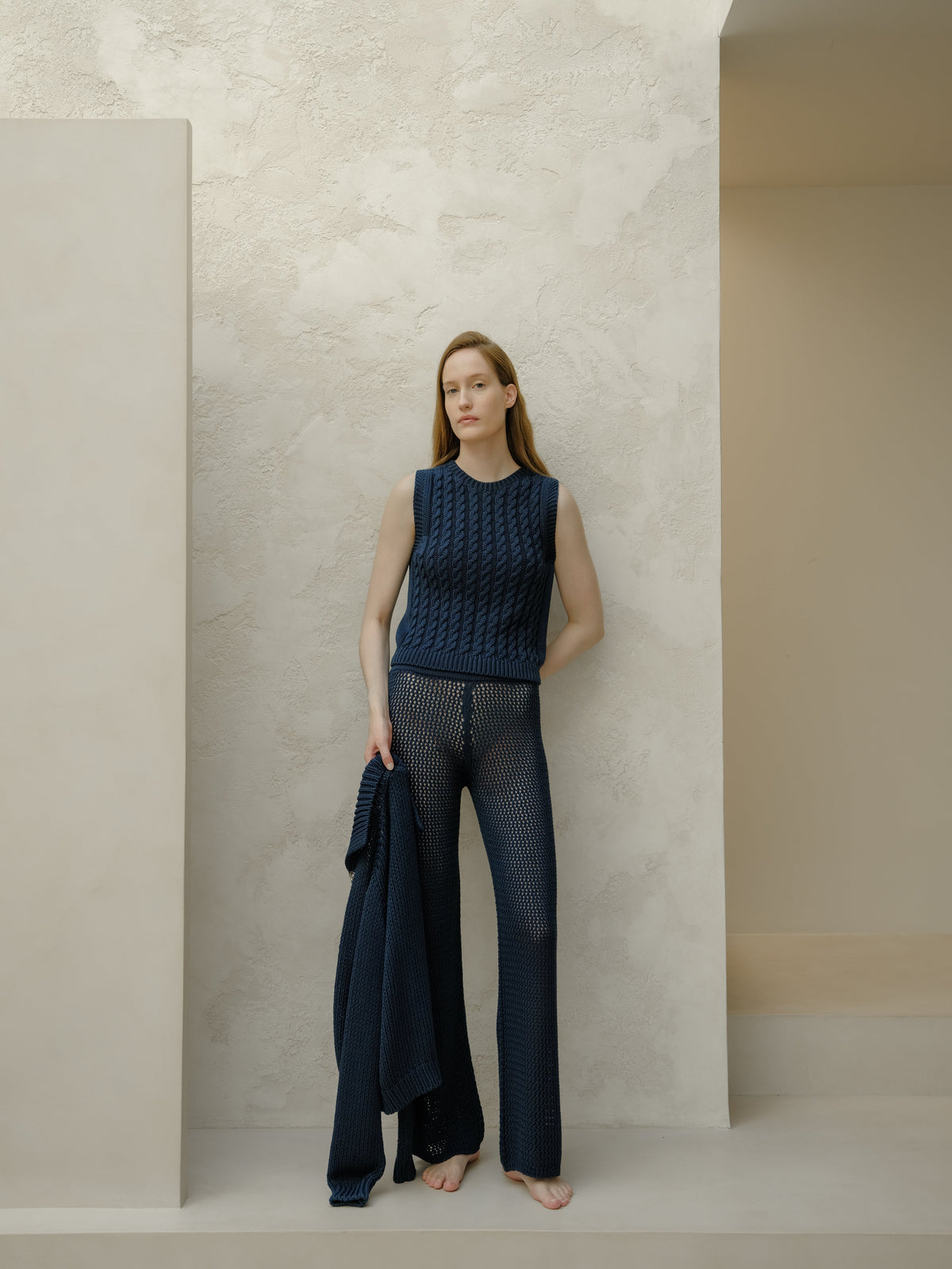 Model in navy knit sleeveless top and navy openwork pants holds a matching navy cardigan against a pale textured wall