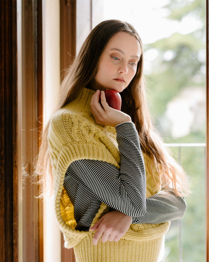 Modella con a crema cashmere mix maxi tunica a trecce over a striped top, holding a rosso apple near her face.
