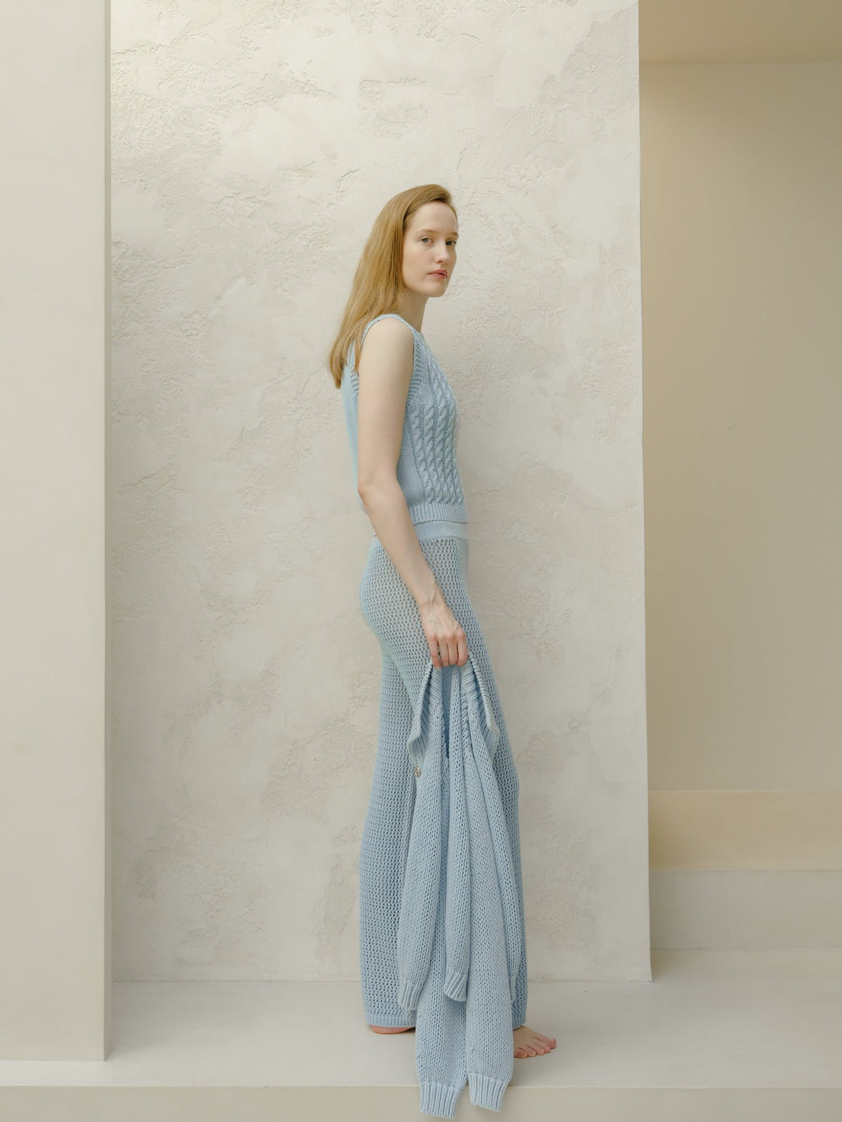 Full-length side view of a woman wearing a light blue sleeveless knit top and matching mesh-knit pants against a pale textured backdrop.